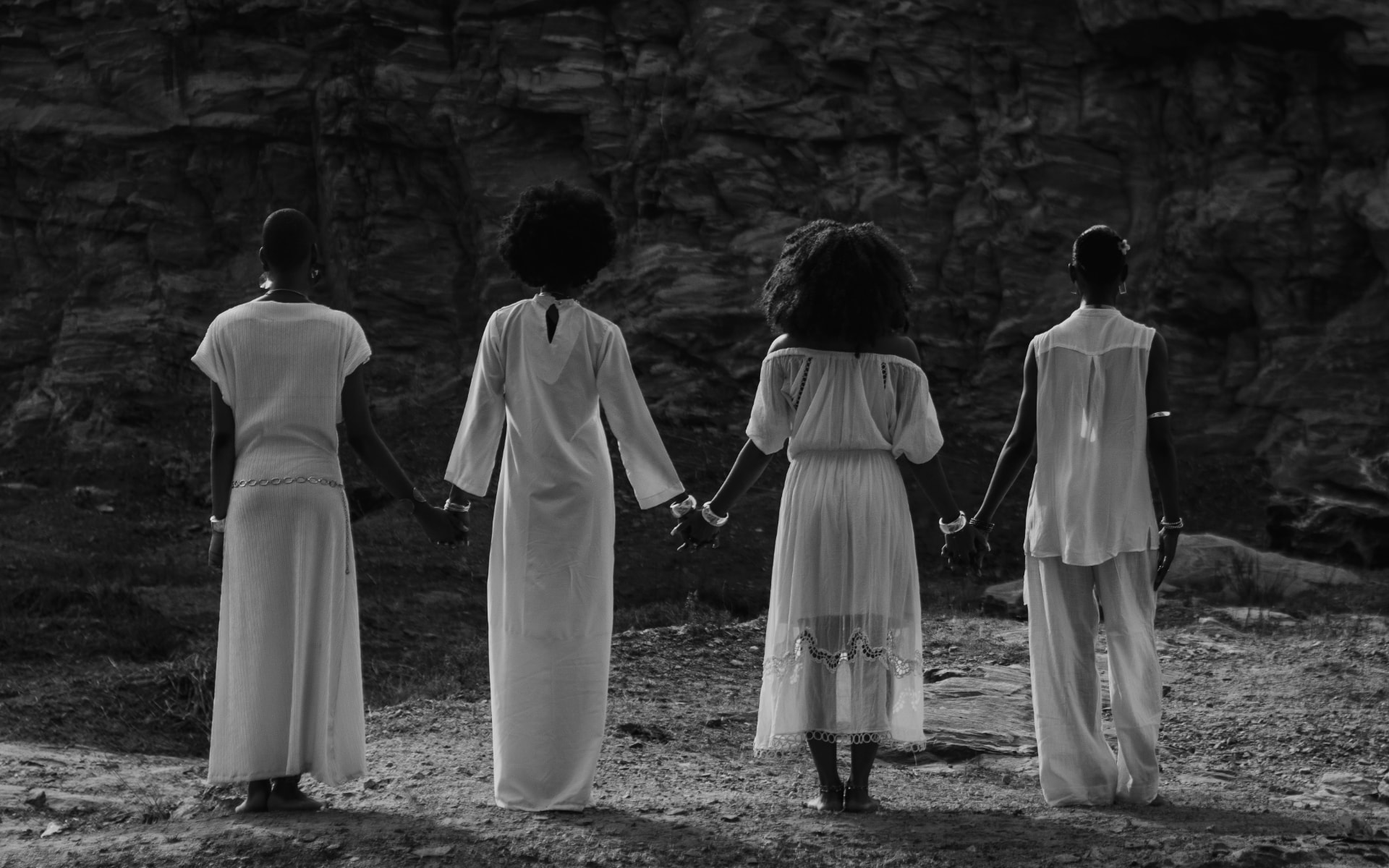 Four women in white dresses holding hands, facing a rocky landscape